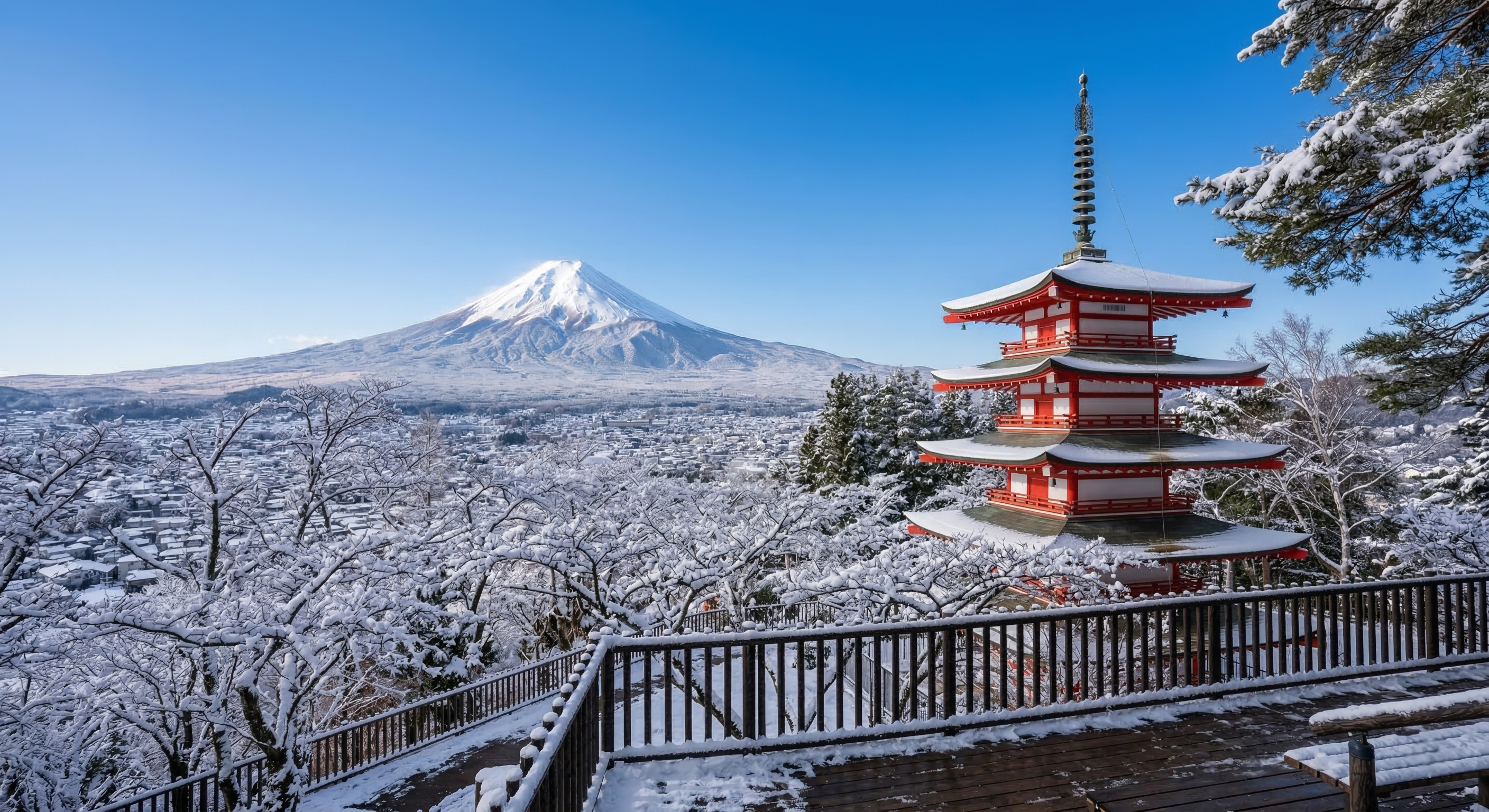 A vibrant red five-story pagoda surrounded by snow-dusted trees overlooks a massive, clear, snow-covered Mount Fuji under a bright blue winter sky during the early morning hours.