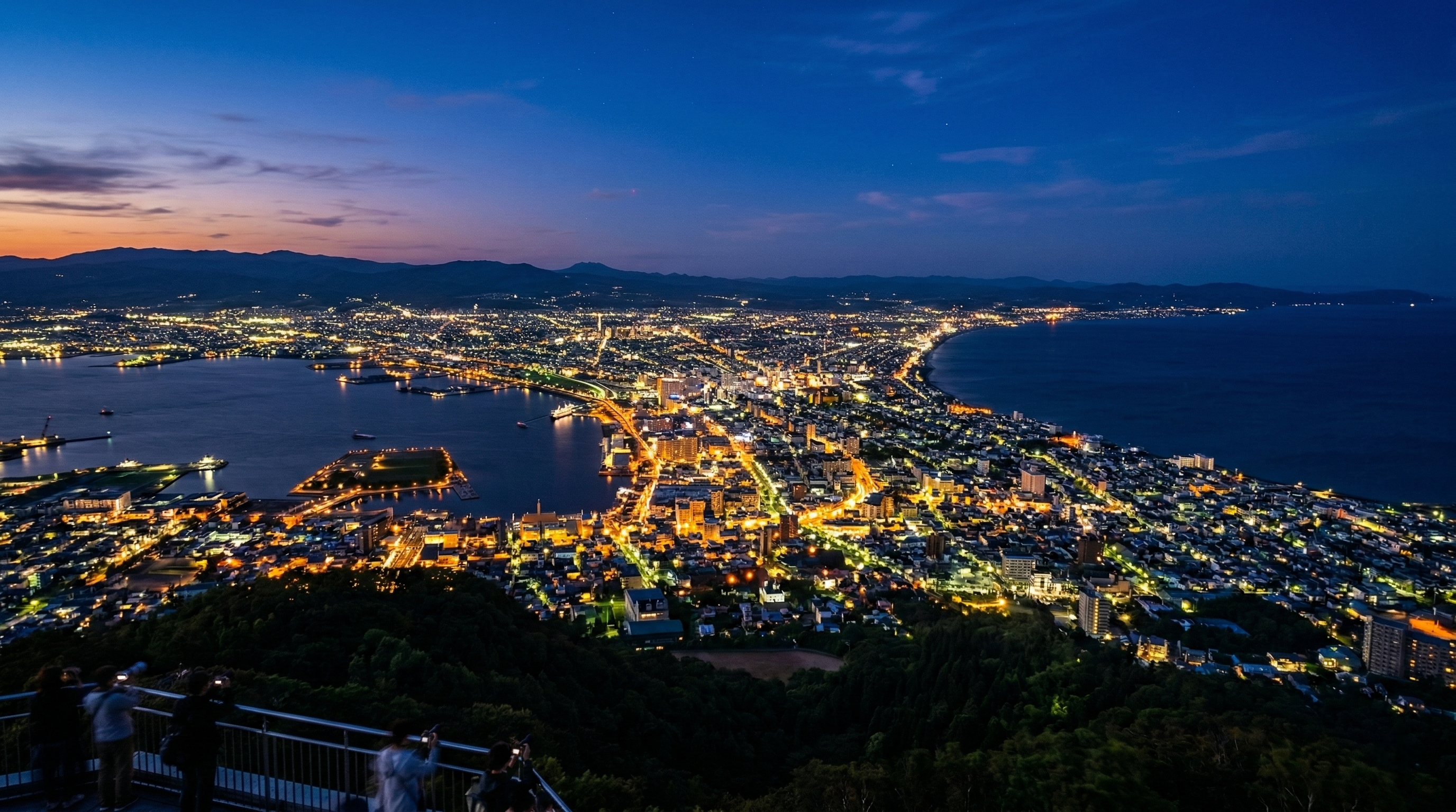 A stunning high-angle view of Hakodate at dusk, showing the narrow urban strip illuminated by golden lights between two dark sea bays under a deep blue sky.