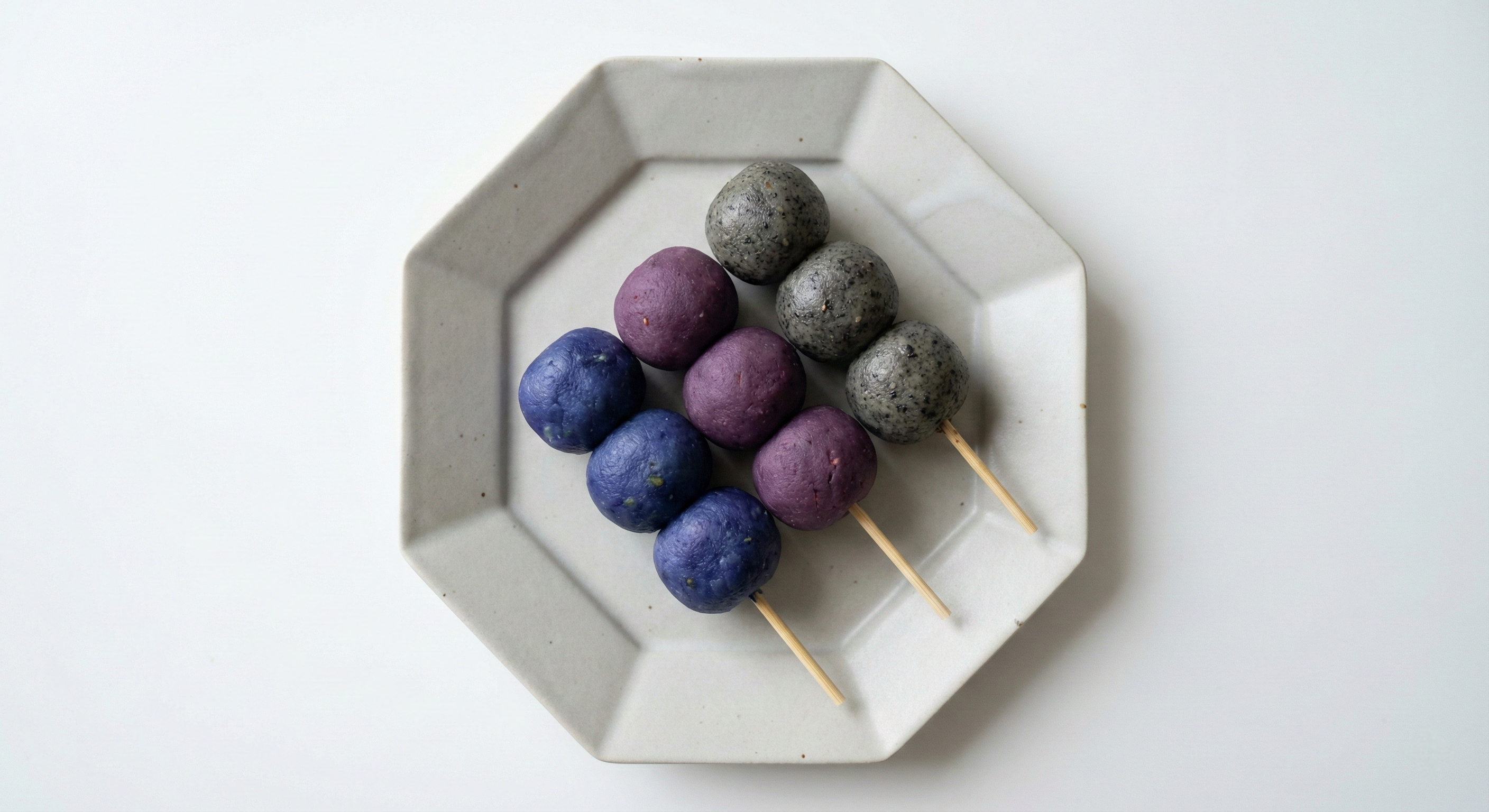 A top-down view of dango skewers in unconventional colors like blue butterfly pea, purple sweet potato, and black sesame, elegantly arranged on a sleek, minimalist gray ceramic plate.