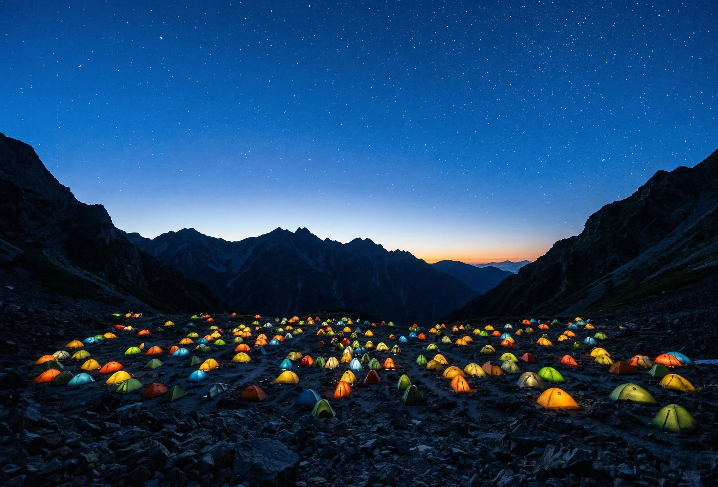 A stunning night shot of numerous illuminated tents glowing within the rocky Karasawa Cirque under a vast, star-filled sky and dark mountain silhouettes.